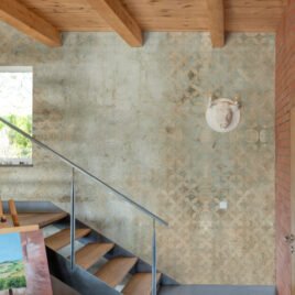 Carta da parati moderna denominata Palinsesto che riproduce una parete materica usurata dal tempo, dove antichi decori geometrici affiorano in trasparenza sotto strati di intonaco grezzo.