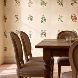 Calda e accogliente cucina rustica di una casa di campagna, con grande tavolo in legno, camino in pietra e pareti rivestite dalla carta da parati naturale L'Erbario Ritrovato.